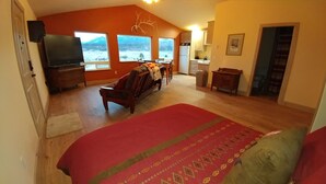 A bedroom with a large bed, a TV, a desk, and a chair. There are windows with a view of mountains.