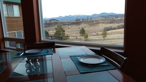 A dining table with two chairs, a glass top, and a view of mountains through a large window.