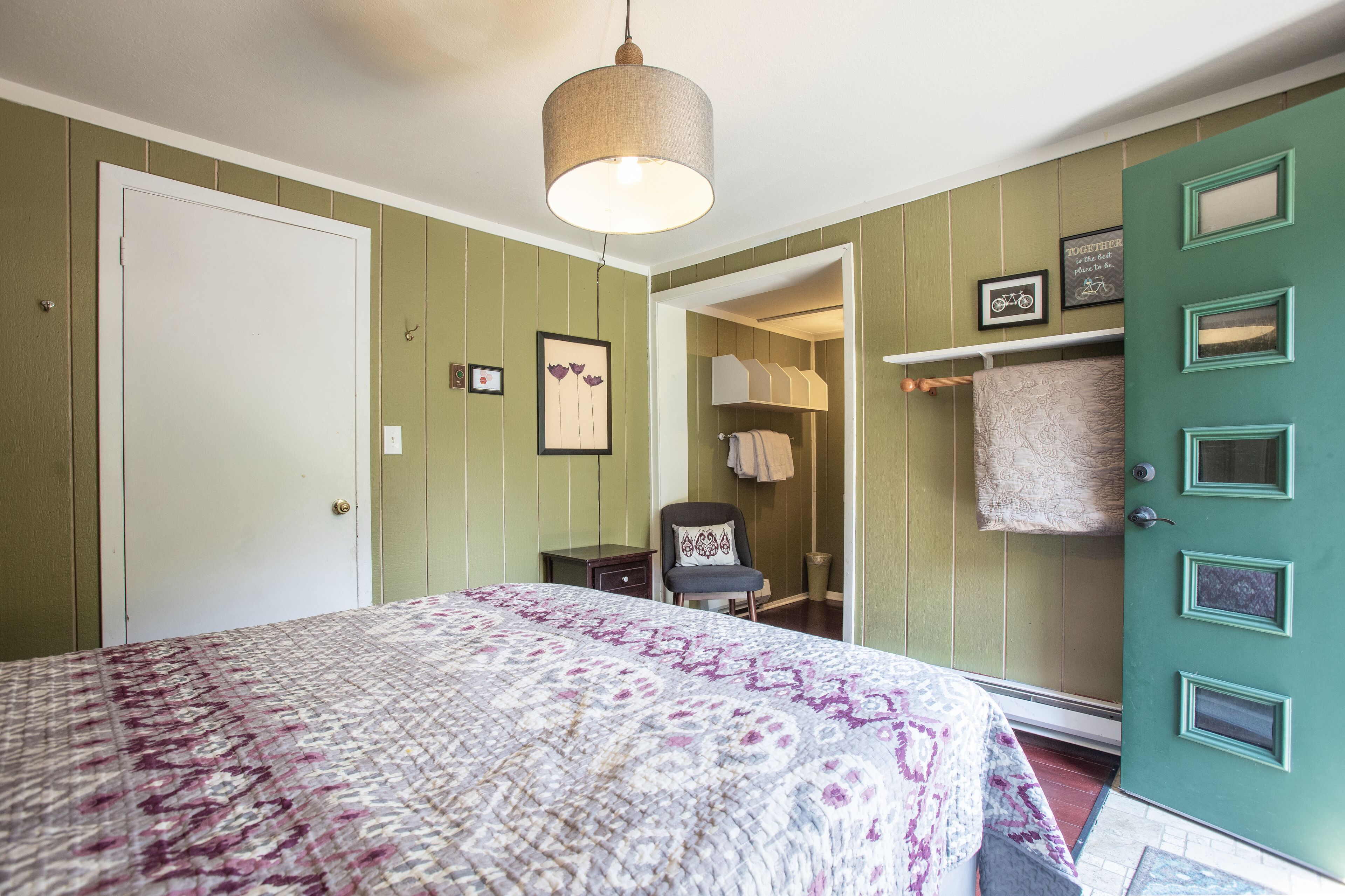 A bedroom with a bed, a chair, a door, and a hanging light fixture.