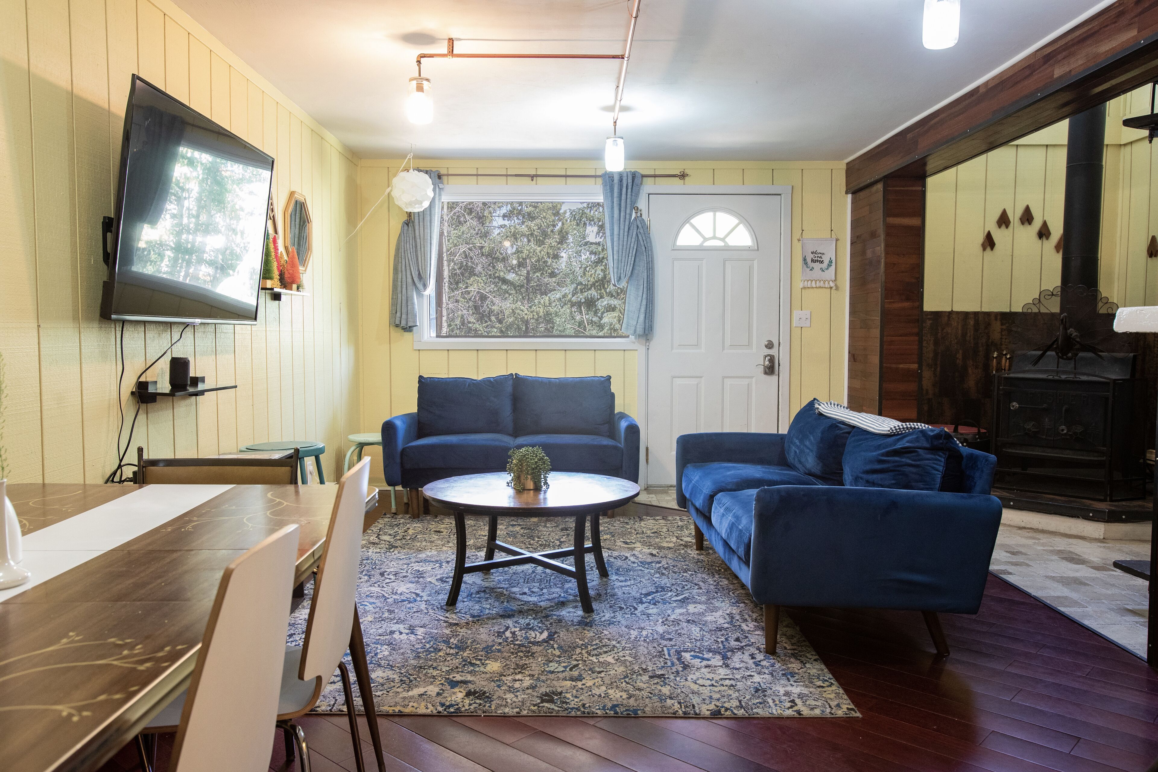 Una sala de estar con una mesa de madera, dos sofás azules, un televisor montado en la pared y una chimenea.