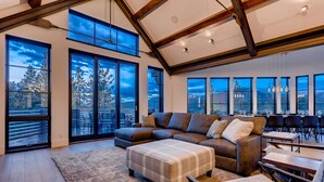 Un salón espacioso con un sofá seccional grande, una mesa de centro y un comedor con mesas y sillas. Amplias ventanas ofrecen vistas al exterior.