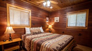 Un dormitorio de cabaña de madera con una cama, dos lámparas de noche, un ventilador de techo y dos ventanas con persianas.