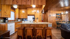Cocina de cabaña de madera con una isla central, taburetes de bar y una zona de comedor con sofá de cuero.