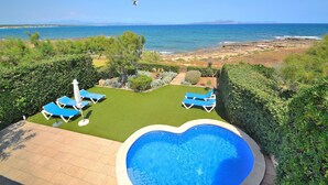 Une piscine dont le bord est en forme de cœur, entourée d’une pelouse avec des chaises longues bleues et offrant une vue sur la mer.