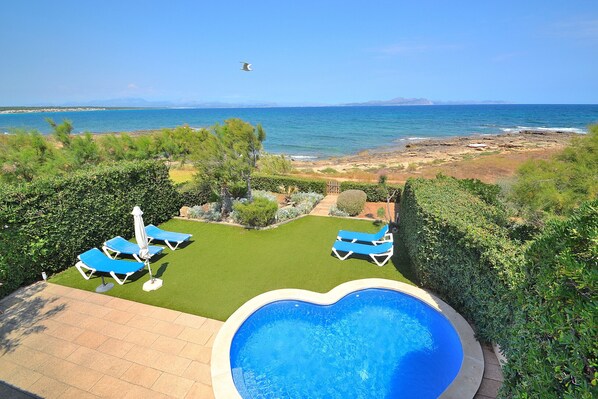Une piscine dont le bord est en forme de cœur, entourée d’une pelouse avec des chaises longues bleues et offrant une vue sur la mer.