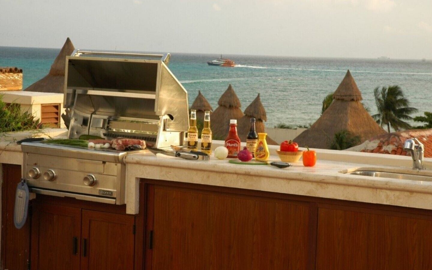 Cozinha externa com churrasqueira, utensílios de cozinha e temperos. Vista para o oceano e um barco ao fundo.