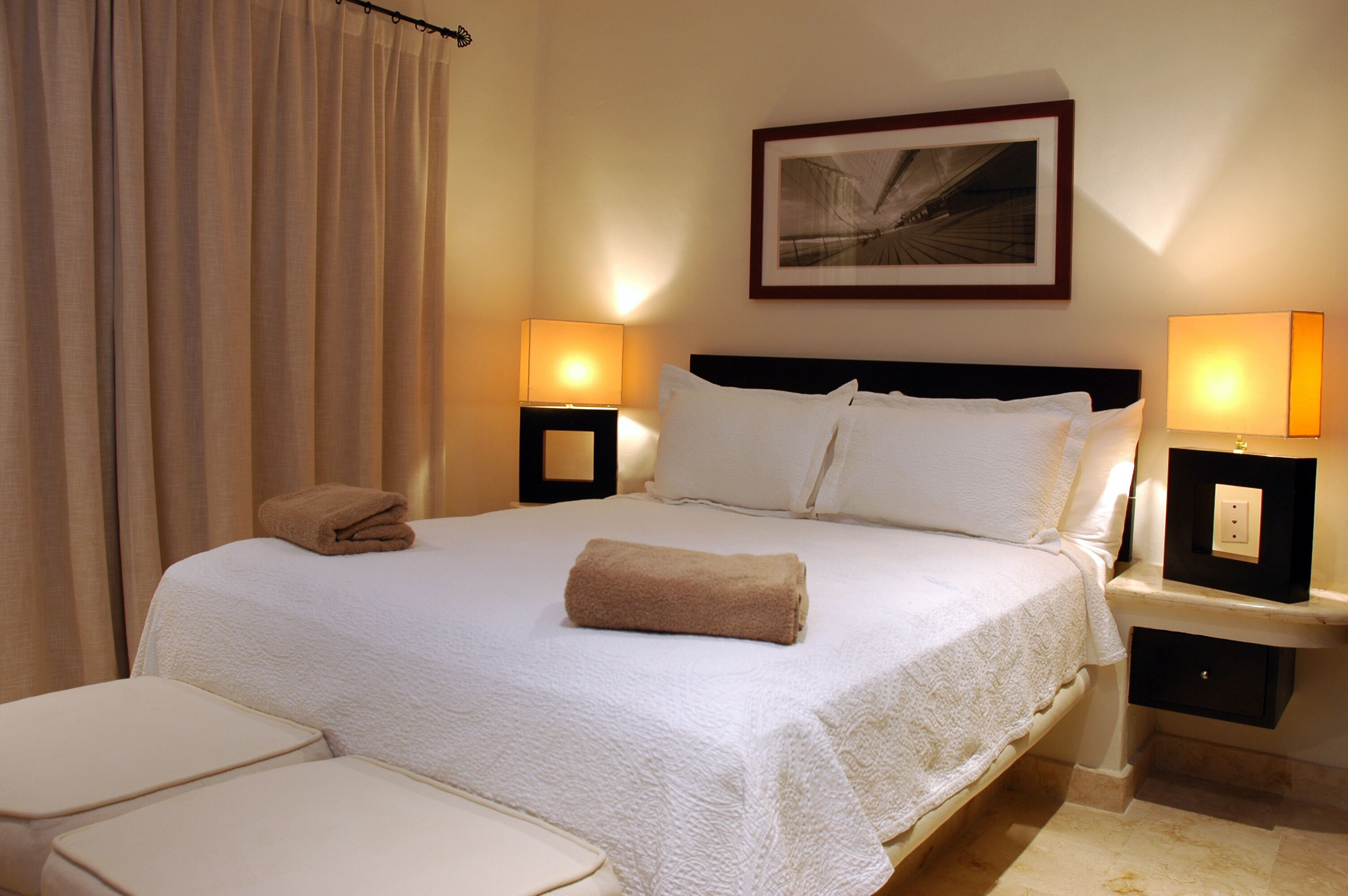 Camera d'albergo con un letto grande, due comodini con lampade, un quadro appeso al muro e una finestra con tende.