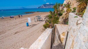 Uma praia com uma escadaria de pedra que desce até a costa, uma placa de sinalização e palmeiras.