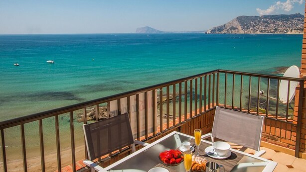 Varanda com mesa posta para o café da manhã, com vista para um mar azul e uma montanha ao longe.