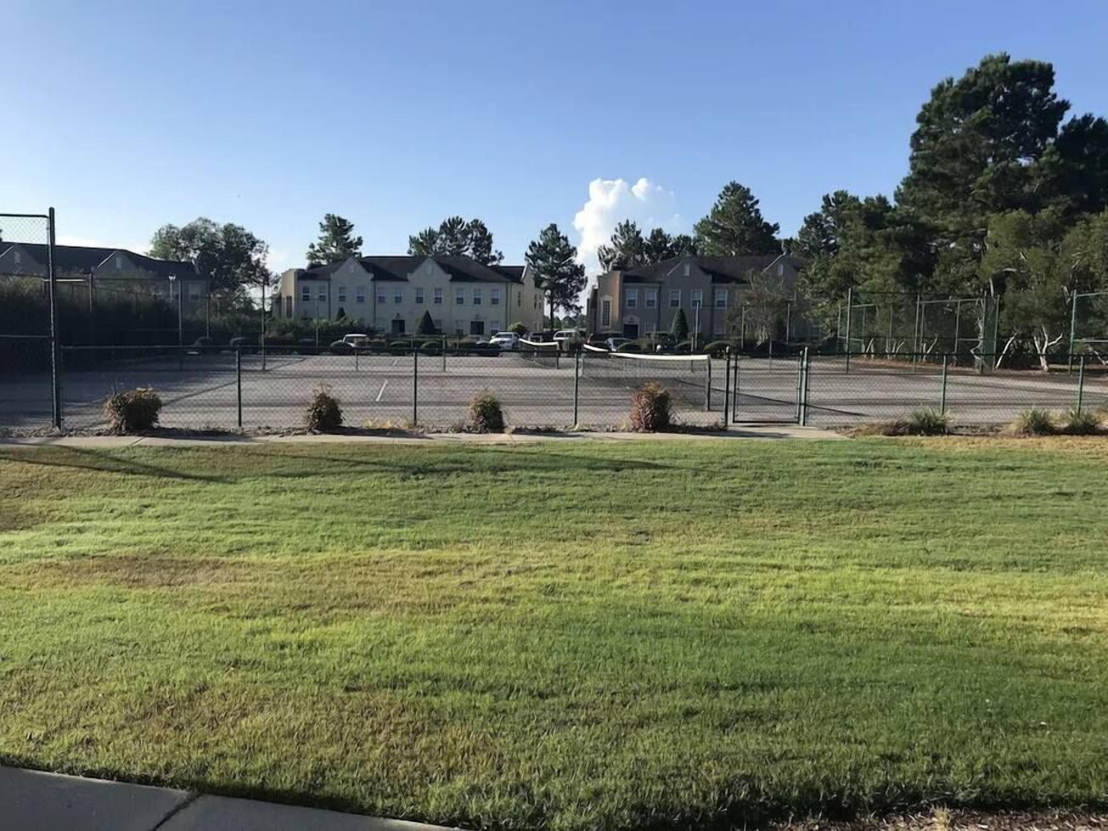 Cancha deportiva cercada con red, rodeada de césped y arbustos, con edificios residenciales al fondo.