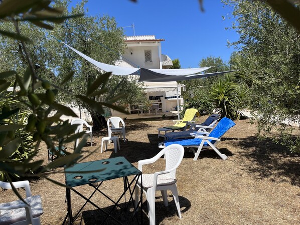 Un patio amueblado con mobiliario exterior, incluyendo sillones reclinables y una mesa, rodeado de follaje y un edificio al fondo.