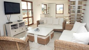 Ein geräumiges Wohnzimmer mit einem weißen Sofa, Korbstühlen, einem weißen Couchtisch und einem an der Wand montierten Fernseher.