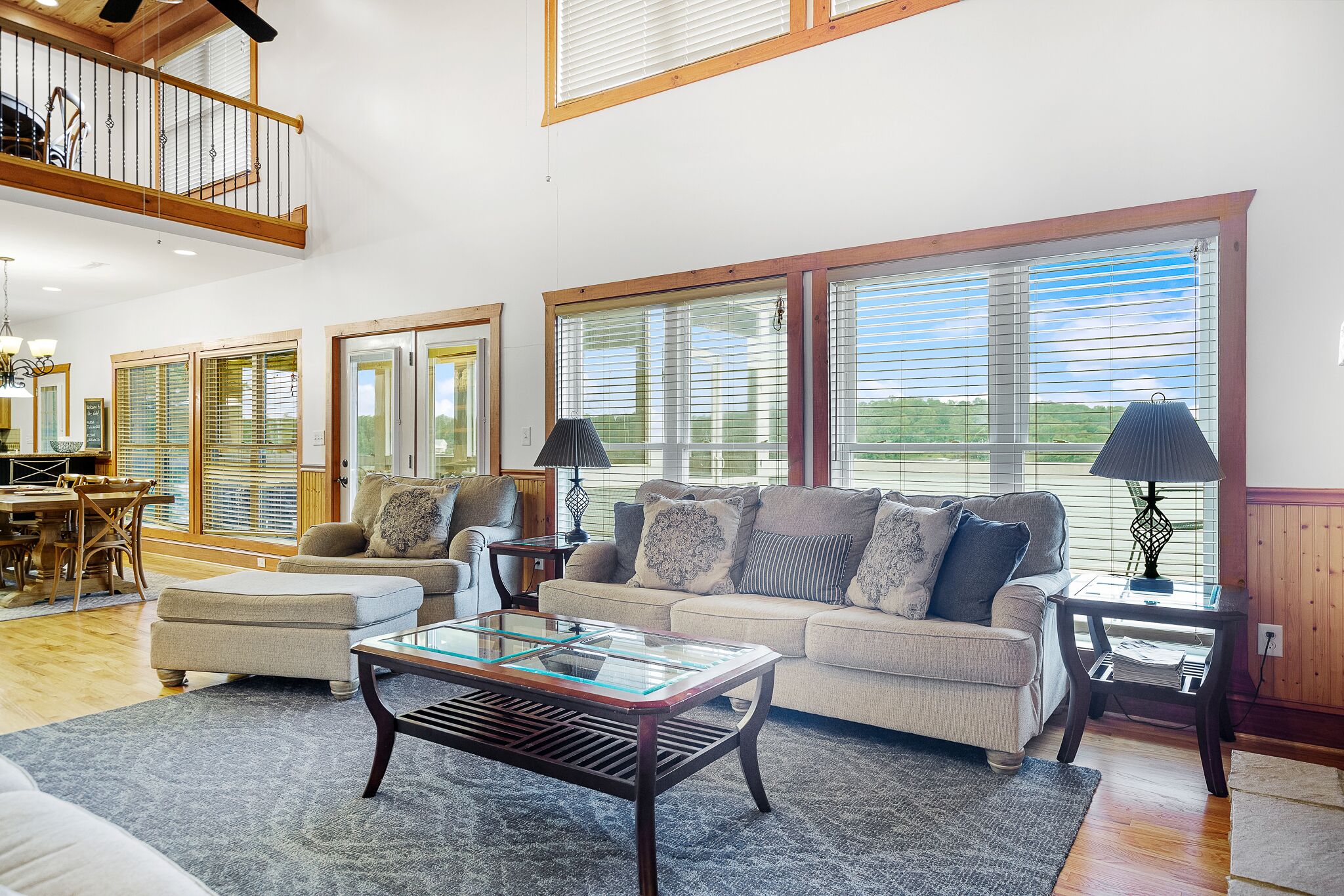 A spacious living room with a high ceiling, large windows, a dining area, and a staircase.