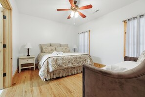 Une chambre avec un plancher en bois, un lit recouvert d’une literie à motifs floraux, un ventilateur de plafond et une table de chevet avec une lampe.