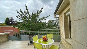 Une terrasse avec une table ronde et des chaises jaunes, un panier de fruits, et une vue sur les arbres et les bâtiments en arrière-plan.