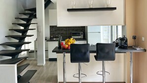 Une cuisine moderne dotée d’un îlot central, d’un four encastré et d’une machine à café. On y trouve deux tabourets de bar noirs et un luminaire suspendu au-dessus de l’îlot.