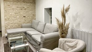 Un salon moderne avec un mur en pierre, un canapé d’angle beige, une table basse en verre et une plante décorative.