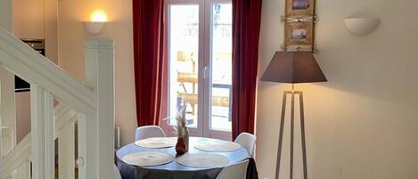 A dining area with a table set for four, a standing lamp, and a view of the outdoors through a door with red curtains.