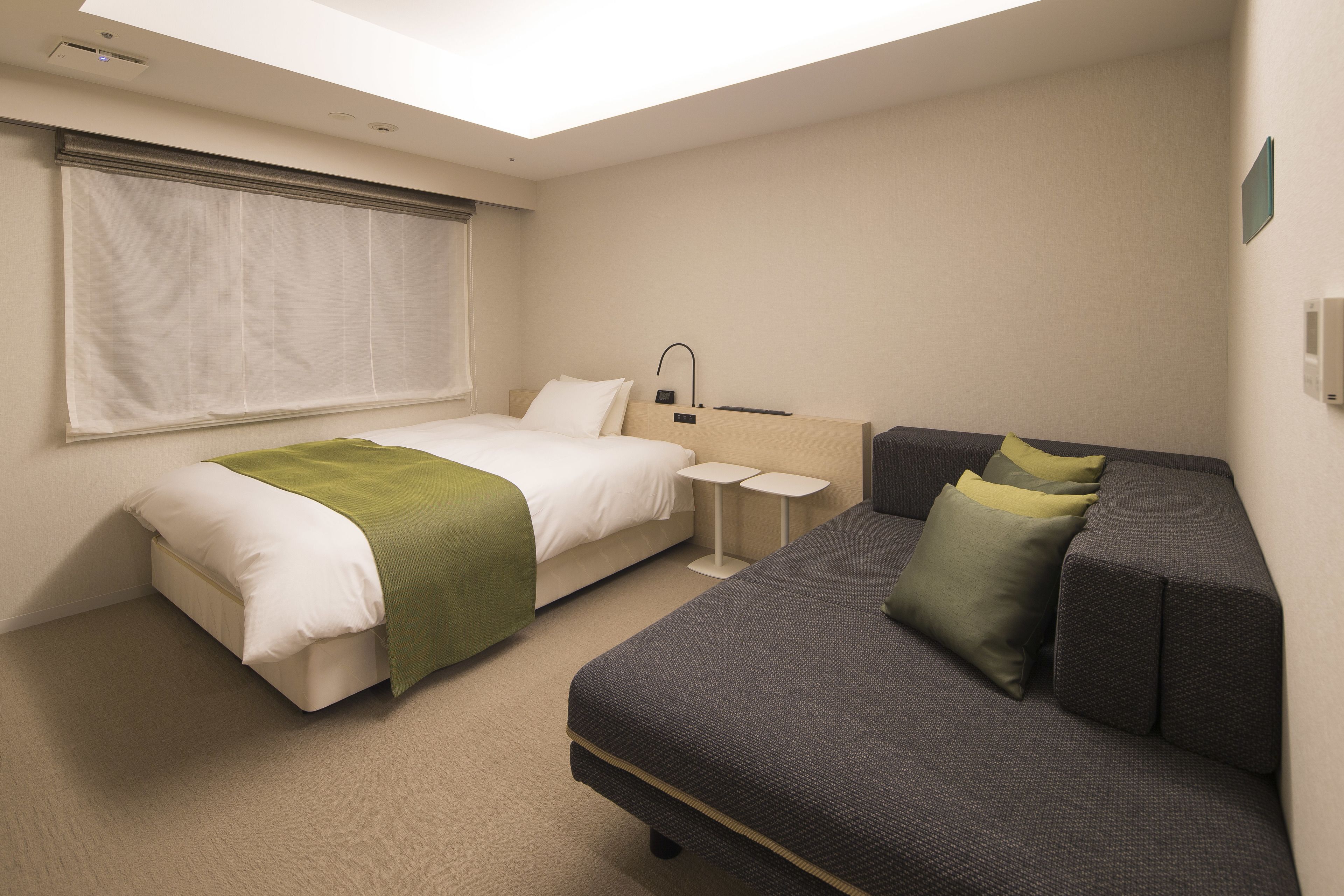 A hotel room with a bed, a sofa, a small table, and a window with blinds.