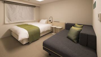 A hotel room with a bed, a sofa, a small table, and a window with blinds.