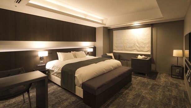 A modern hotel room with a large bed, bedside tables, a desk, and a bench.