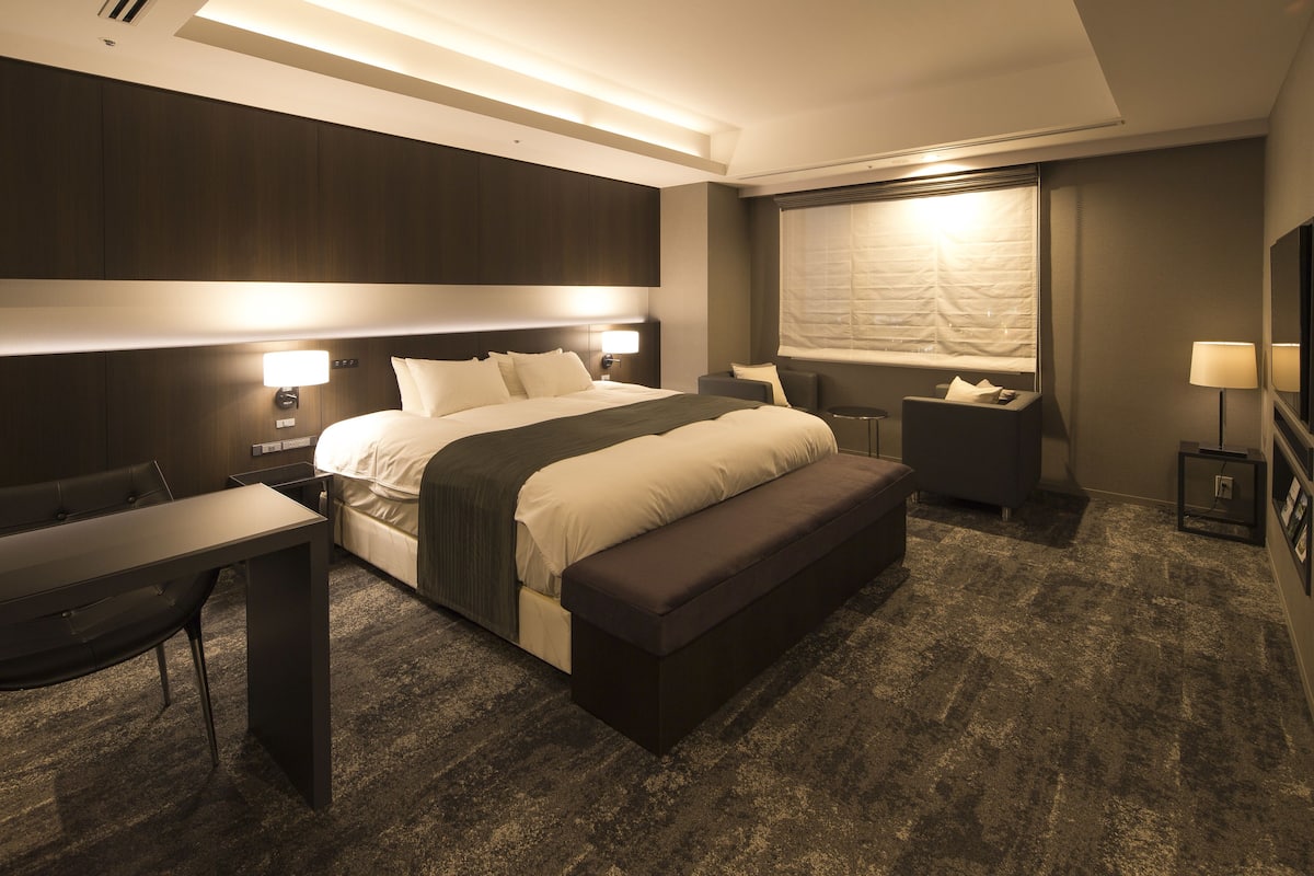A modern hotel room with a large bed, bedside tables, a desk, and a bench.