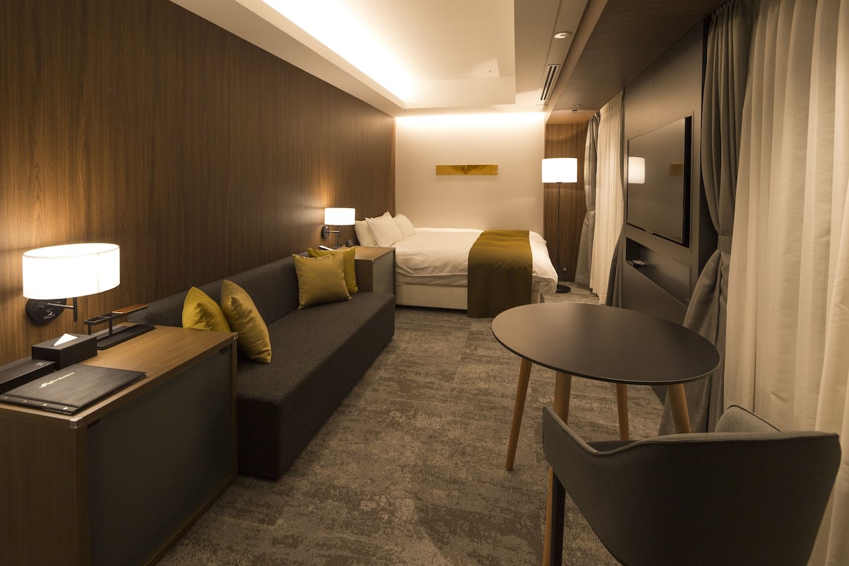 A hotel room with a bed, a sofa, a desk with a lamp, a chair, and a TV.