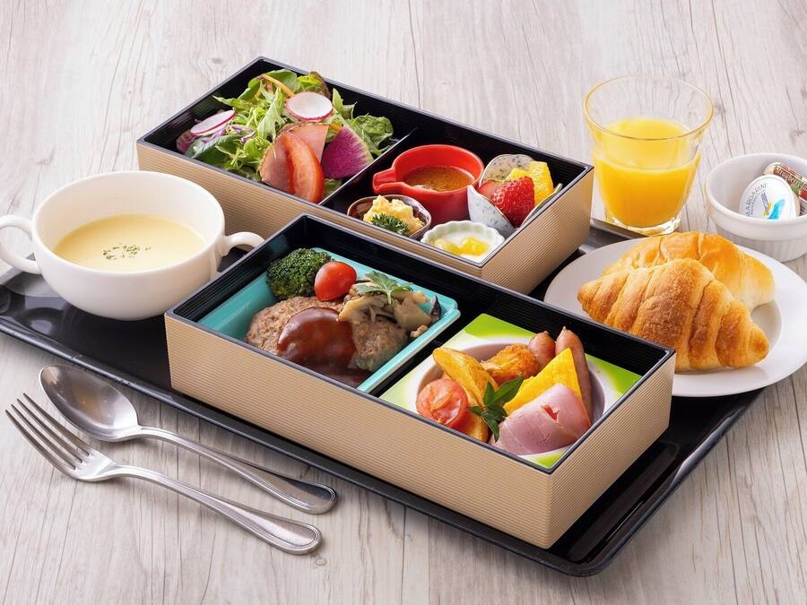 Eine Schale mit Bento-Boxen, ein Glas Orangensaft, ein Croissant und eine Suppentasse.