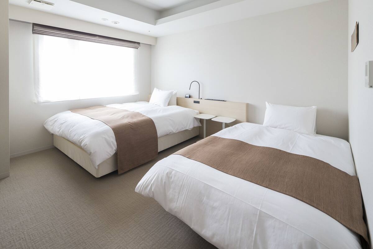 A hotel room with two beds, a bedside table, a lamp, and a window with blinds.