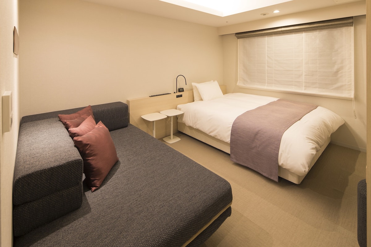 A hotel room with a sofa, a bed, a small table, and a window with blinds.