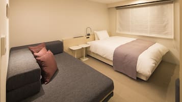 A hotel room with a sofa, a bed, a small table, and a window with blinds.