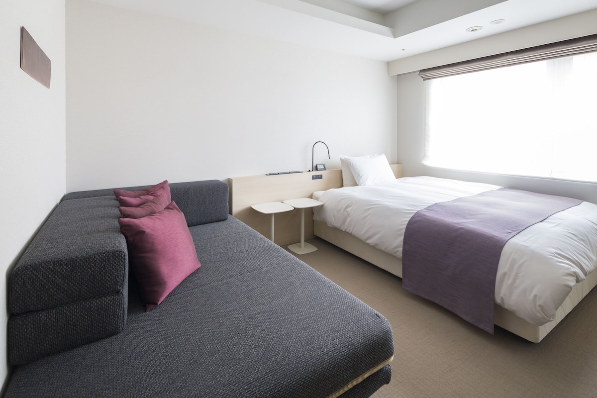 A modern hotel room with a grey sofa, a single bed, and a small bedside table with a lamp.