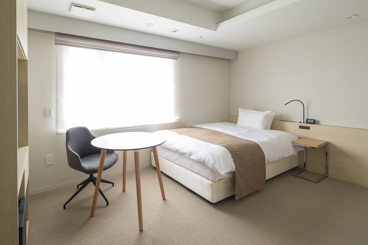 A hotel room with a bed, a small table, a chair, a bedside table, and a window with blinds.