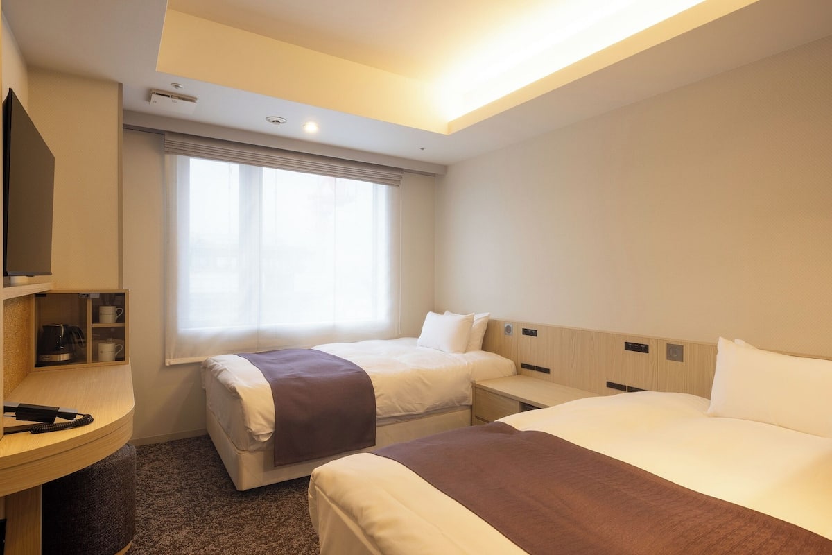 A hotel room with two beds, a desk, a TV, and a window with blinds.