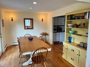 Un comedor con mesa y sillas de madera, una cocina con armarios y electrodomésticos, y una repisa con vajilla.