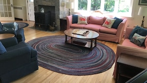 A cozy living room with a stone fireplace, a pink sofa with crab-themed pillows, a blue armchair, and a coffee table with a chess set. - Spacious Cottage Overlooking Bass Harbor with Sweeping Front Lawn (Bass Harbor)