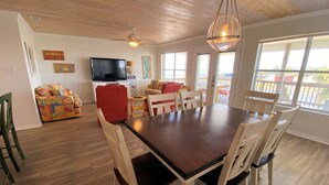 A dining area with a wooden table and chairs, a television, and artwork on the walls. - Sunset Key - Soundfront with Boat Dock, 3 Bedrooms, 2 Baths, Sleeps 9. (Dauphin Island)