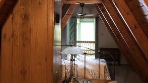 Une chambre confortable située sous les toits, avec des poutres en bois, un ventilateur de plafond, une table de chevet agrémentée d’un vase, et une vue sur le balcon extérieur.