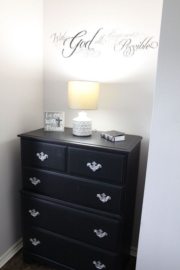A black dresser with silver handles, a white wall with a motivational quote, a white lamp, and a small framed picture.