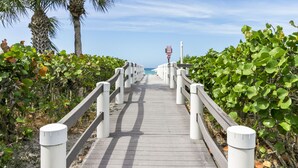 A wooden boardwalk with white railings, leading towards a body of water, surrounded by greenery.