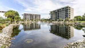 A modern building with multiple floors and balconies, surrounded by a calm water body and greenery.