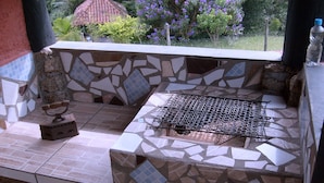 Un área de parrillada con mosaicos, una parrilla, un taburete de madera y una funda metálica para la parrilla.