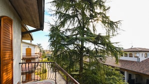En balkon med jerngitter, der ud til en gårdsplads med et stort træ og en bygning med et tagtækket tag.