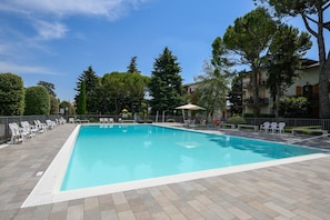 Una piscina rettangolare con bordo piastrellato, circondata da sedie e ombrelloni bianchi, con alberi e un edificio sullo sfondo.