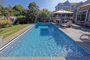 Una piscina rectangular con borde de azulejos, rodeada por un patio con sillones de descanso y una zona de estar cubierta.