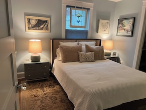 A bedroom with a bed, two bedside tables with lamps, a window with a view, and artwork on the walls.