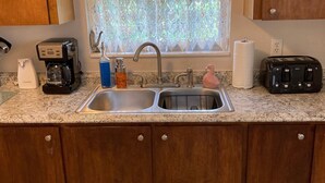 A kitchen with a double sink, wooden cabinets, a window with lace curtains, and a toaster. - CHARLOTTE HARBOR BUNGALOW (Port Charlotte)