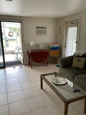 Uma sala de estar com uma mesa de centro de vidro, um sofá, uma mesa lateral de madeira, uma cadeira vermelha e uma porta que dá para o exterior.