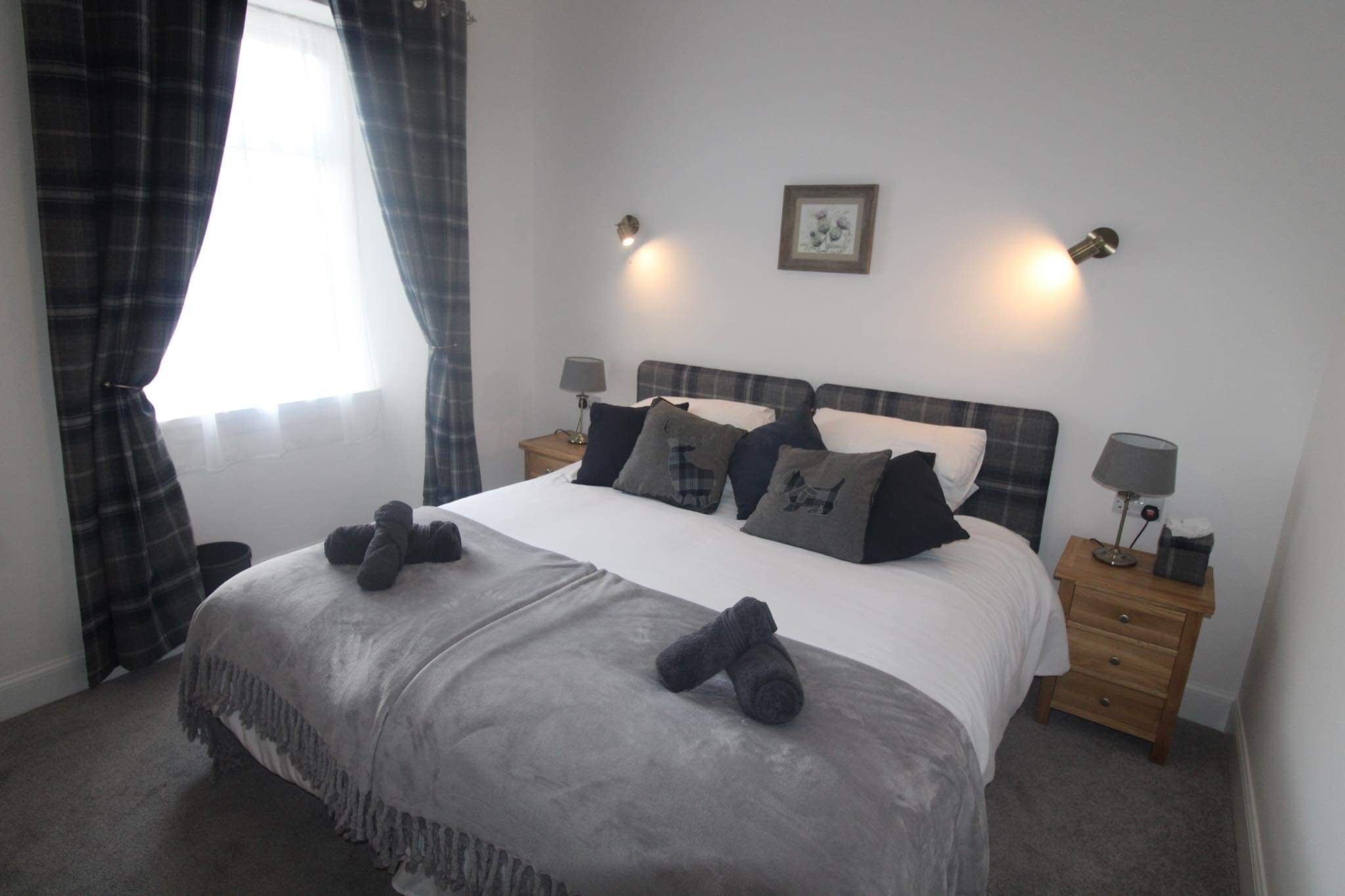 A neatly made bed with a grey blanket and two rolled-up towels, a bedside table with a lamp, and a window with curtains.