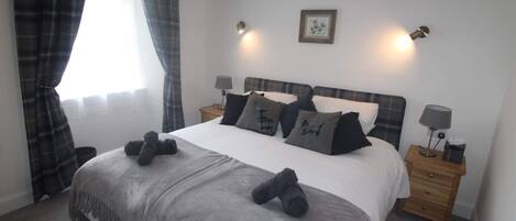 A neatly made bed with a grey blanket and two rolled-up towels, a bedside table with a lamp, and a window with curtains.